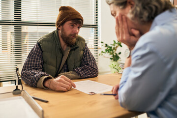 Middle aged Caucasian man sitting across from middle aged Caucasian woman social worker at desk discussing documents during social services meeting