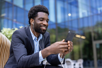 A smiling businessman in a suit is looking at his phone while wearing earphones, outdoors near a modern building. He appears relaxed.