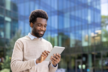 Plakat A smiling businessman looks at his tablet while standing outdoors near a modern office building.