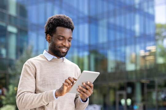 Fototapeta A smiling businessman looks at his tablet while standing outdoors near a modern office building.