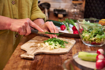 Woman chopping green onions on a wooden cutting board in the kitchen, close-up of hands