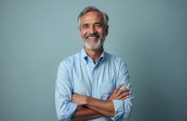 Happy mature Indian man with crossed arms smiles. Gray hair, dressed in blue shirt. Positive emotion, well-being, success. Studio portrait on plain background, business or finance theme, retirement.