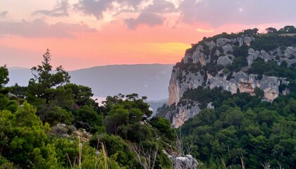 Fototapeta premium Scenic view of rocky mountain cliff with green trees under a colorful sunset sky in the distance