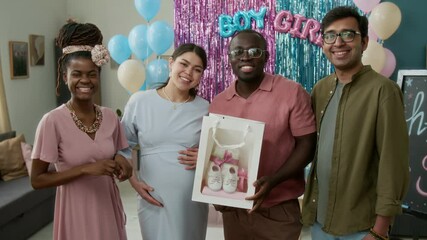 Medium full portrait of happy diverse expecting couple with multiethnic friends standing in living room with pink, blue decorations at gender reveal party, holding new baby hamper, smiling at camera
