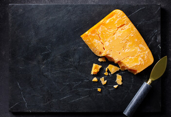 Aged cheddar cheese with knife on black marble serving board. Dark background. Copy space. Top view.