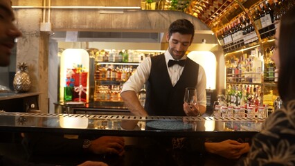 Focusing smiling smart bartender serving gin fizz or tonic soda at luxurious bar, engaging lively conversation with VIP customer couple at back view in atmosphere at lighting warm nightclub. Vinosity.
