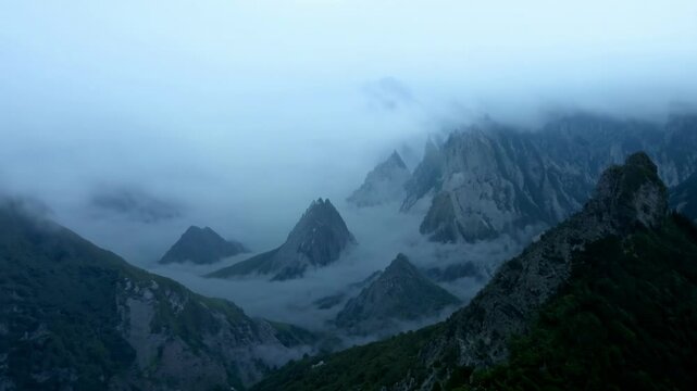 Majestic mountain peaks emerge through thick rolling fog and mist creating ethereal atmospheric landscape with dramatic weather conditions and moody blue-gray tones in natural wilderness setting