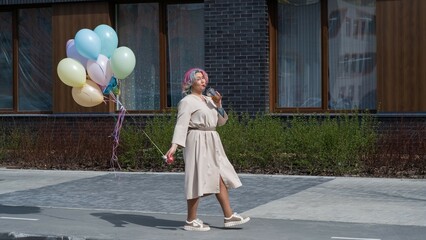 Woman in colored hair walks with an armful of balloons and drinks a refreshing beverage
