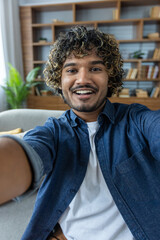 A smiling young man takes a selfie with a friendly expression while seated at home. His curly hair...