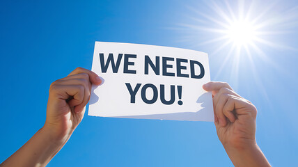 Hands holding a sign that says we need you against a blue sky with bright sun