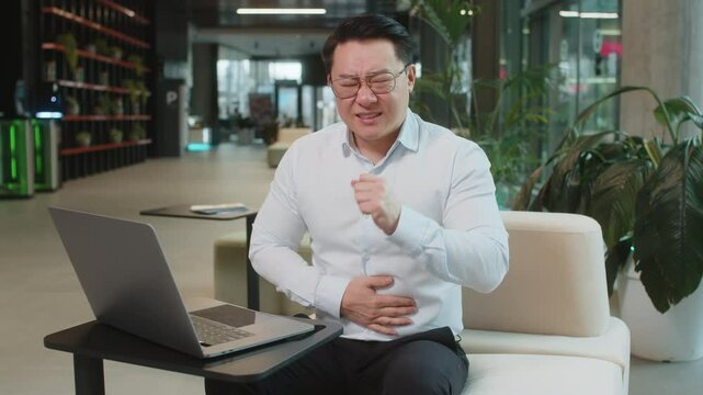 Disappointed sick Chinese businessman feeling overeating during work process on laptop computer in office lobby. Upset ill male manager guy suffering from painful stomach ache period cramps, poisoning