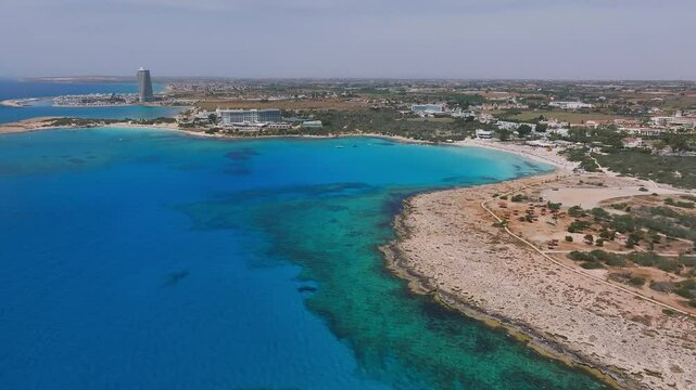 Aerial pan of Ayia Napa, Cyprus, showing turquoise waters, vibrant beaches, beachfront resorts, a modern tower, and lush greenery.