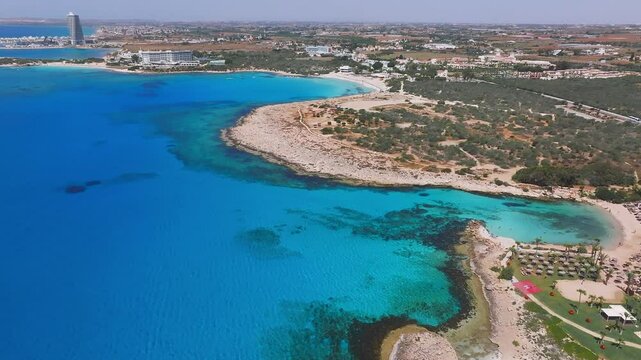 Aerial view of Ayia Napa, Cyprus, showing turquoise waters, rocky shores, Love Arch, resorts, Nissi Beach, Landa Beach, and gentle wave motion.