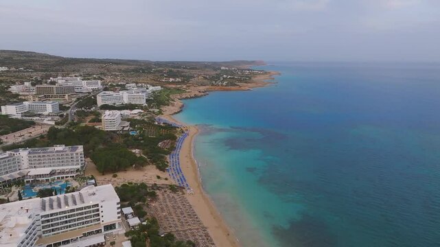 Aerial view of Ayia Napa, Cyprus, showcasing turquoise waters, sandy beaches, resorts, rocky outcrops, and colorful umbrellas along the shore.