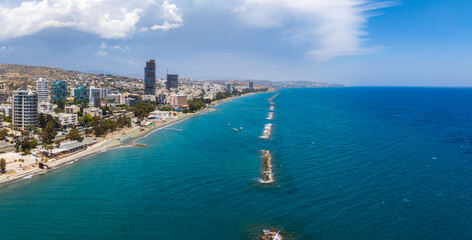 Fototapeta premium Aerial view of Limassol, Cyprus, showing modern high rises, sandy beaches, promenades, clear blue sea with breakwaters, and rolling hills under clouds.