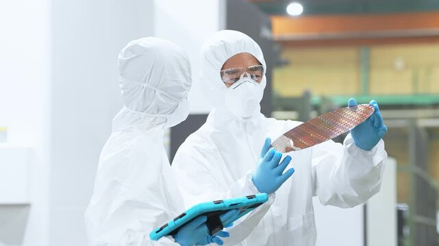 半導体工場でタブレットを持ってシリコンウエハーを検査する防護服を着た男女作業員