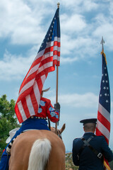 A woman in a blue outfit adorned with floral embroidery rides a horse, holding the United States flag.