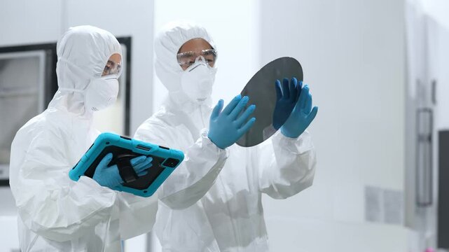 半導体工場でタブレットを見ながらシリコンウエハーを検査する防護服を着た男女作業員