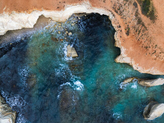 Aerial view of Cyprus coastline with turquoise waters, white limestone cliffs, rocky outcrops, pebbled shoreline, and reddish brown terrain above.