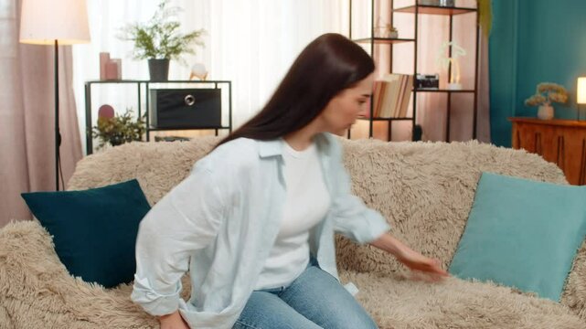 Young confused woman at home anxiously searching for lost apartment keys between sofa pillows exhaling relief after finding them. Caucasian girl stressed about losing object expressing joyful reaction