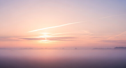 Naklejka premium Soft hues of pink and orange fill the sky as the sun rises above the horizon, casting light on a foggy landscape. The tranquil scene evokes a sense of peace and solitude in the early morning.
