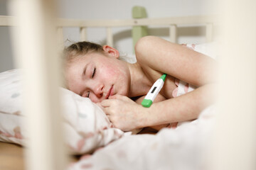 A young girl sleeping in a bed, appearing unwell. She is resting on a pillow with a thermometer in her hand, suggesting she may be sick. The scene is intimate and conveys a sense of care and concern.