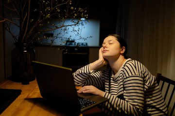 A young woman with a cell phone in hand and a laptop sits at the dining room table in her kitchen...