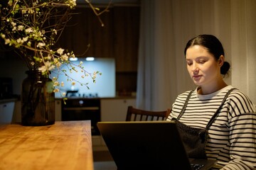 A young woman with a cell phone in hand and a laptop sits at the dining room table in her kitchen...