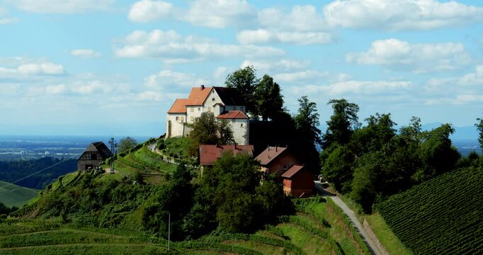 Aerial drone footage slowly zooms out from Schloss Staufenberg castle in Durbach, revealing red-roofed buildings, green vineyards, and scenic hills under a sky filled with fluffy clouds and sunlight