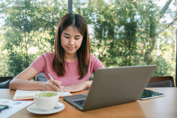 A young woman in a pink shirt works on her laptop and takes notes in a cozy cafe surrounded by lush greenery.