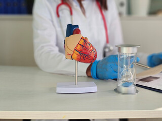 A healthcare worker in coat inspects a heart model on a table, holding a notepad. An hourglass sits...
