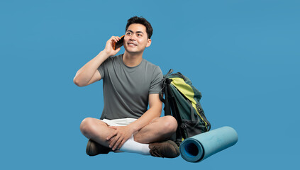 Smiling hiker chats on phone sitting cross-legged with backpack and mat against blue backdrop
