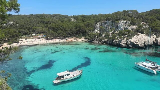 Crystal-clear turquoise waters of Cala Macarella beach in Menorca, surrounded by pine trees and rocky cliffs, with boats anchored in the peaceful bay.