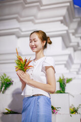 asian woman in traditional dress holding a flower arrangement and wai gesture during the khan dok flower offering festival at wat chedi luang in chiang mai, thailand.

