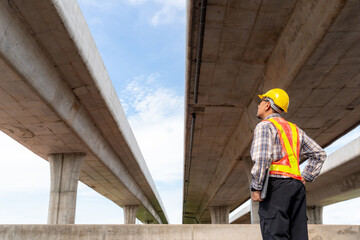 An engineer, wearing a protective helmet and safety vest, inspects the structural integrity of a concrete overpass against a clear, bright sky.