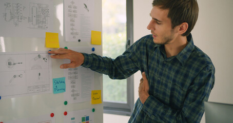 Male engineer points to technical drawings and planning notes on a whiteboard during a robotics project presentation in a modern tech lab.