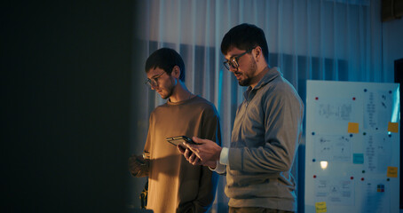 Caucasian engineers discuss project strategy using digital tablet in a modern tech office at night....