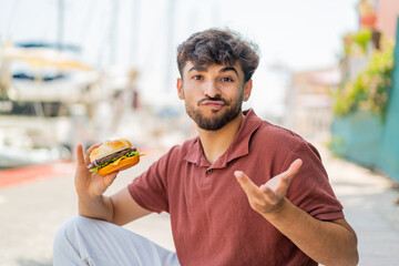 Young Arabian handsome man holding a burger at outdoors making doubts gesture while lifting the...