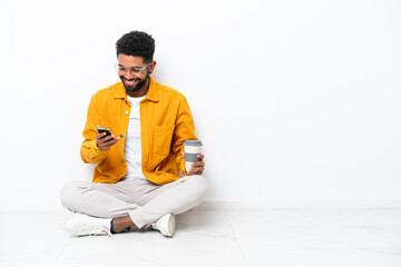 Young Brazilian man sitting on the floor isolated on white background holding coffee to take away and a mobile