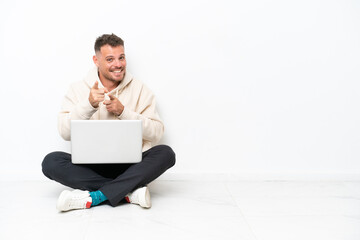 Young caucasian man with a laptop sitting on the floor isolated on white background points finger at you with a confident expression
