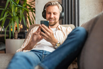 Attractive young man relaxing on a couch at home, listening music and playing on smartphone.