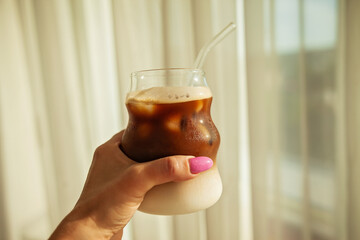 A woman's hand holding a homemade iced coffee latte by the window, bathed in soft morning light -...