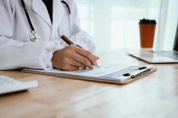 Fototapeta premium Focused female doctor working at her desk in a hospital, using a laptop and clipboard for patient consultation and diagnosis