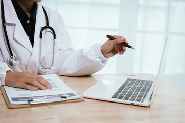 Fototapeta premium Focused female doctor working at her desk in a hospital, using a laptop and clipboard for patient consultation and diagnosis