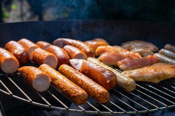 Grill loaded with sausages and marinated chicken, sizzling over hot charcoal, with smoke rising in the sunlight.