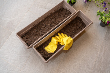Two empty planters filled with moist soil stand on the balcony floor, with yellow gardening gloves...