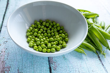 pile of empty green pea pods next to white bowl of shelled peas on rustic wooden table side view