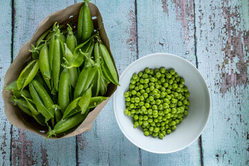 white bowl of shelled green peas next to paper bag of empty pea pods on rustic wooden table, top view