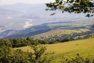 Fototapeta premium Panoramic view of a traditional village nestled among green rolling hills, with a winding river and distant mountains under a hazy sky. Perfect for travel and landscape.