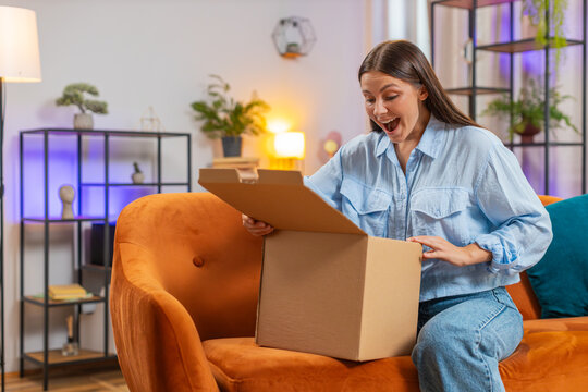 Happy Caucasian young woman unpacking delivery parcel sitting on home couch. Girl shopper online shop customer opening cardboard box receiving purchase gift by fast postal shipping at room apartment.